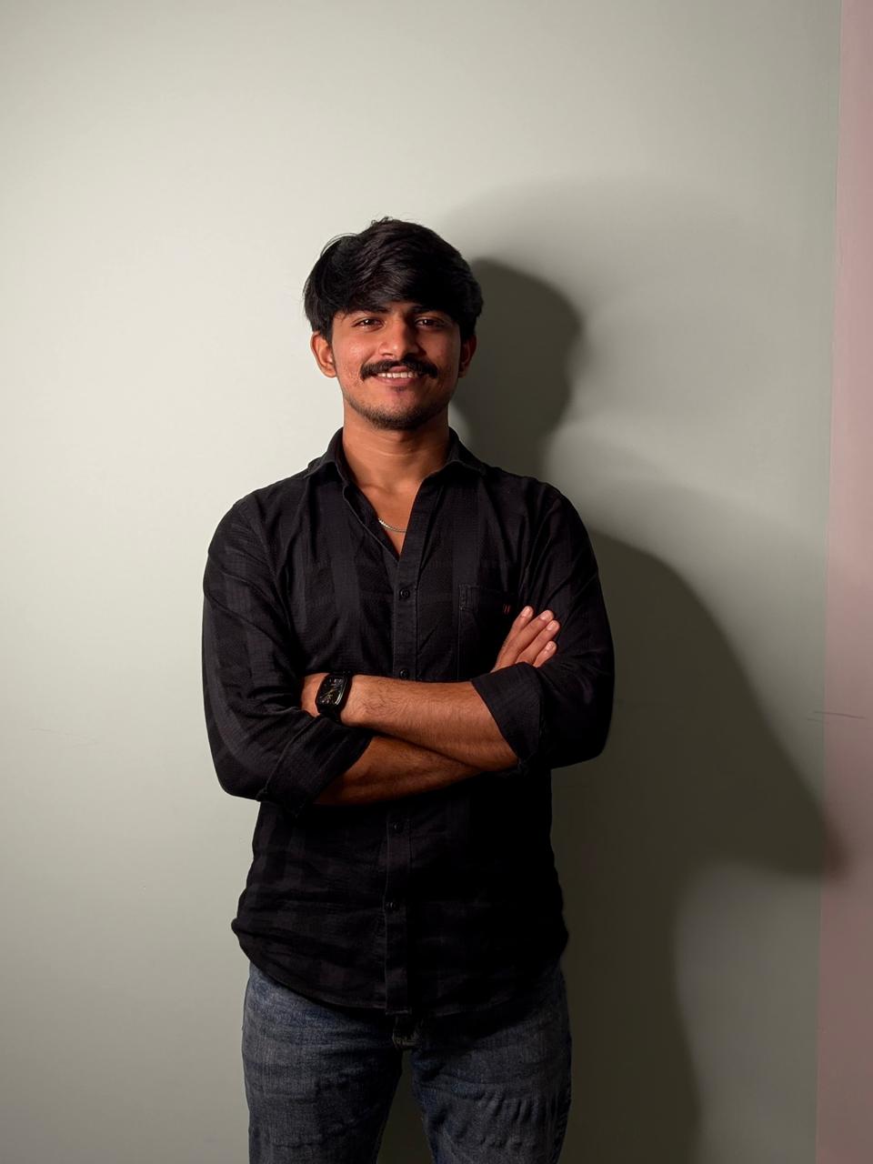 “Man standing with arms crossed, wearing a black shirt, smiling confidently against a plain light background.”